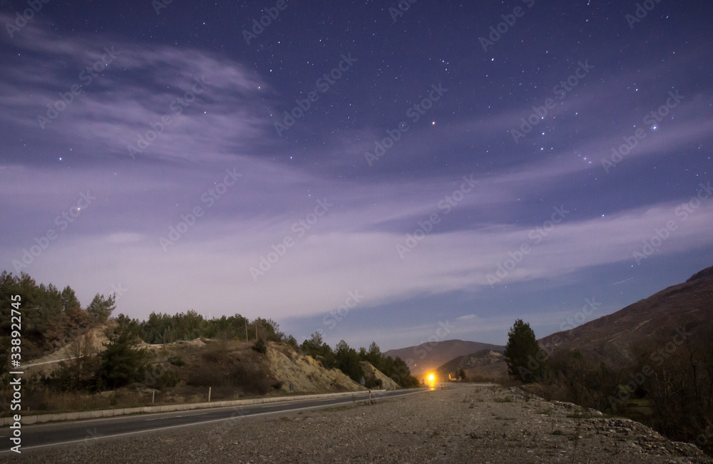 Long exposure photo of purple sky night with stars and clouds, road and yellow light