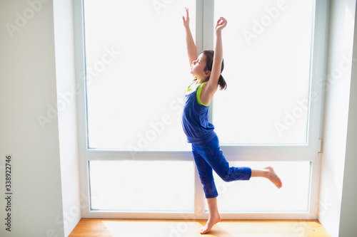 Fototapeta Naklejka Na Ścianę i Meble -  Little girl reaching up workout at home. Cute kid is training on a wooden windowsill indoor. Little dark-haired female model in sportswear has exercises near the window in her room.