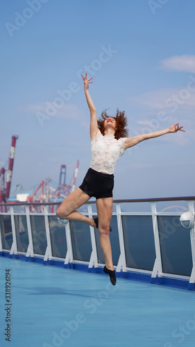 Sporty slim dancer fulfills jumping poses, promenade deck of cruise ship, background cargo port cranes, professional. graceful red-haired curly girl, ballerina. Modern dance choreography, performer