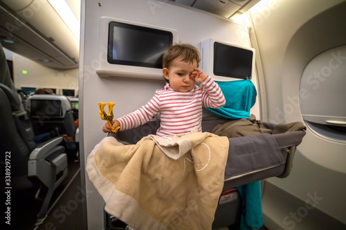 sleepy little cute toddler sitting in the baby bassinet of an airplane with toy rubs his eyes. close-up, soft focus