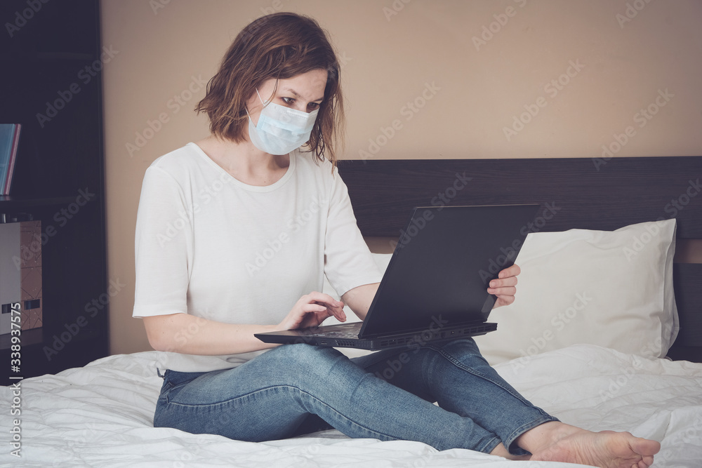 Woman in medical mask working working on a bed with laptop computer. Coronavirus quarantine, isolation period covid - 19. Social distancing