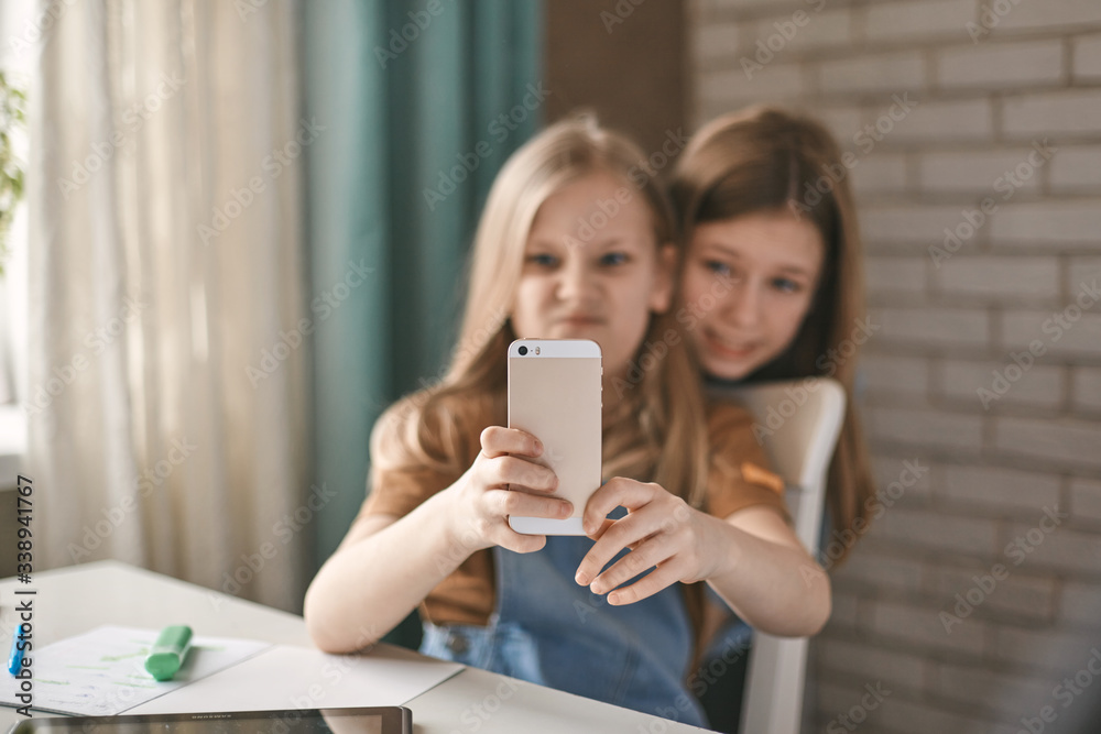 Two sisters take a break from homework and take selfies on their mobile phone while sitting at a table. Education, distance learning, home schooling during quarantine Two sisters take a break from homework and take selfies on their mobile phone while sitting at a table. Education, distance learning, home schooling during quarantine