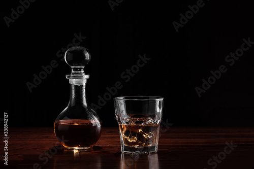 Crystal decanter or carafe of whiskey and glass with alcoholic drink and ice on wooden table in restaurant. Black background for copy space