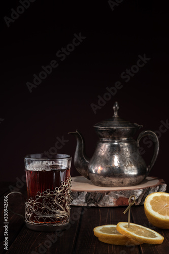 vintage iron kettle with patterns and paintings and a glass of tea with lemon in a cup holder on a wooden table, concept of restaurant menu with copy space