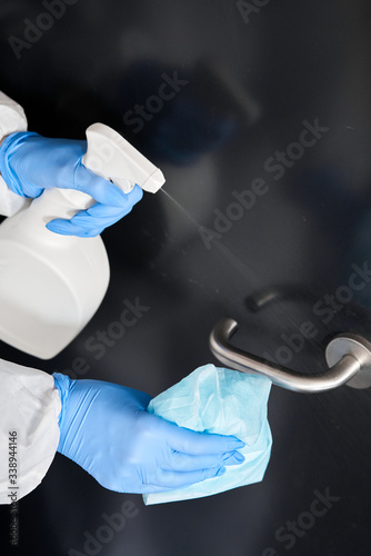 Disinfection, cleaning and washing of door handles. COVID-19. Prevention of coronavirus infection. A woman in a protective suit and blue gloves sprays a disinfectant solution. Prevention of