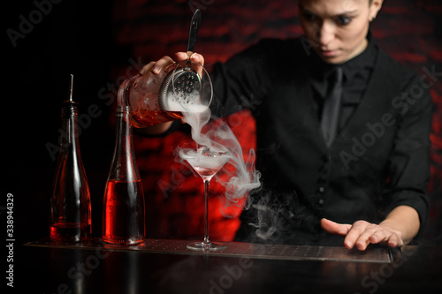young female barman gently pours smoky cocktail from mixing bowl into martini glass.