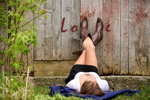 Woman laying on the ground with her cowboy boots on the side of a barn spelling out the word Love. photo