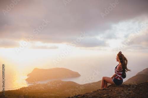 Young woman sitting on a edge of cliff at the sunset, looking at Assos village on Kefalonia island, Greece.