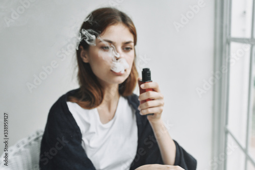 A woman at home in a blanket smokes a vape near the window