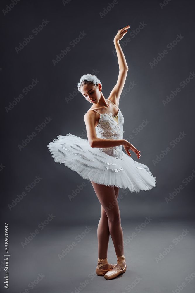 Naklejka premium A young graceful ballerina dressed in professional attire, pointe shoes and a white tutu, demonstrates dance skills. Beautiful classic ballet dancer isolated on grey backgroung in photostudio