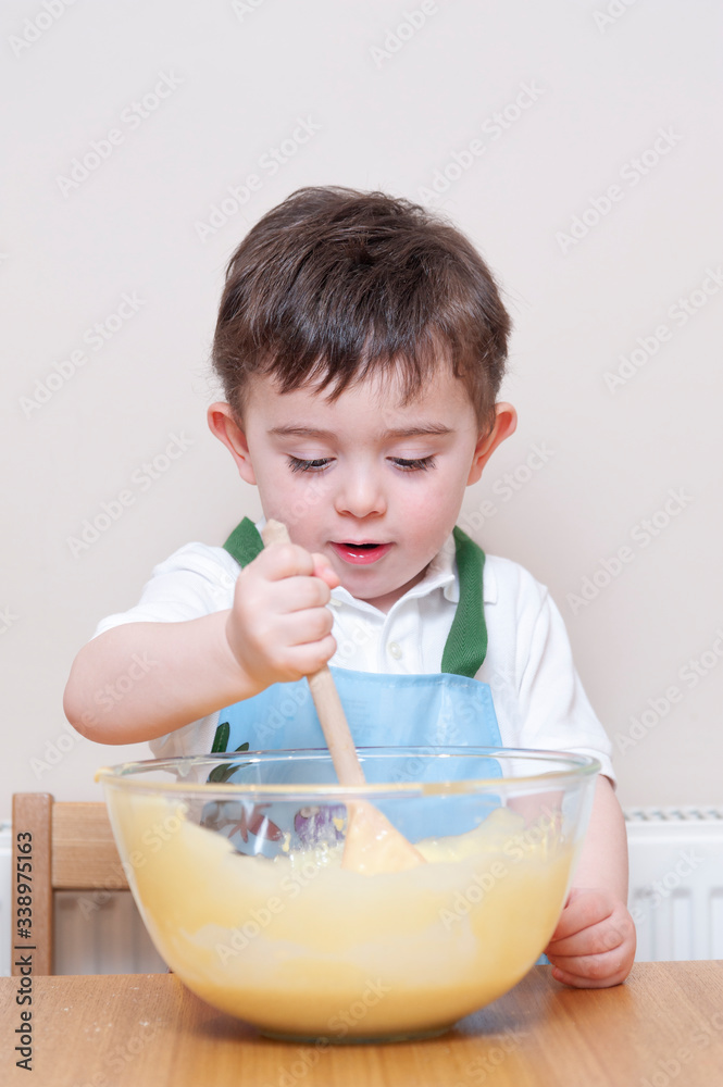 Cute young toddler wearing an apron stirring cake mix inside a glass bowl. He is using a wooden spoon and is sat in his kitchen Cute young toddler wearing an apron stirring cake mix inside a glass bowl. He is using a wooden spoon and is sat in his kitchen
