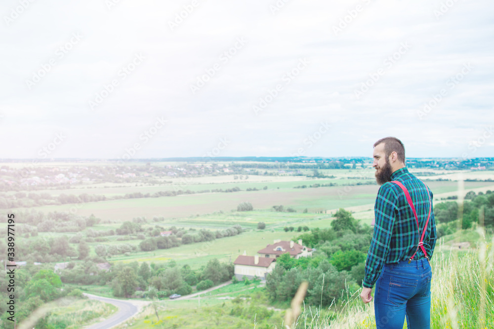 Handsome man hipster with beard on serious face in cloth shirt and suspenders sunny outdoor on mountain top against cloudy sky on natural. Tourism concept. Wonderful landscape.