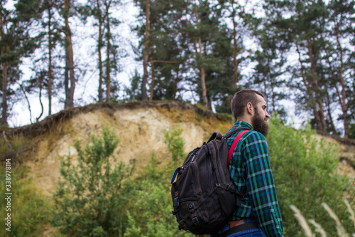 Handsome man hipster with beard on serious face in cloth shirt and suspenders sunny outdoor on mountain top against cloudy sky on natural. Tourism concept. Travel with a backpack. Wonderful landscape.