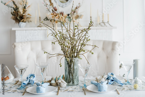 Decorative festive table setting with sky blue textile napkin and floral tablecloth, easter eggs, dishes and golden cutlery, glasses. willow in vase. holiday home interior.