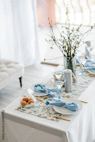 Decorative festive table setting with sky blue textile napkin and floral tablecloth, easter eggs, dishes and golden cutlery, glasses. willow in vase. holiday home interior.