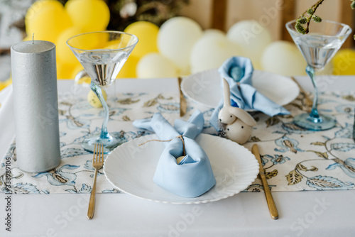 Festive table setting for traditional dinner. sky blue textile napkin, floral tablecloth, easter eggs, golden fork and knife, willow. Yellow balloons party decoration