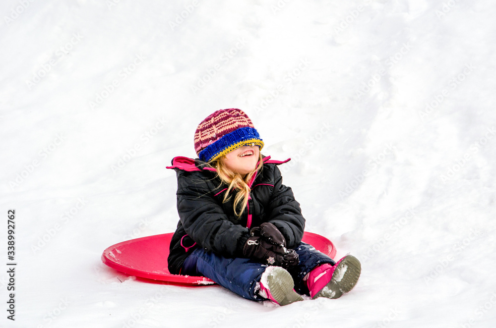 Laughing girl with hat over her eyes, sits on a red saucer sled as she plays on a snowy winter day
