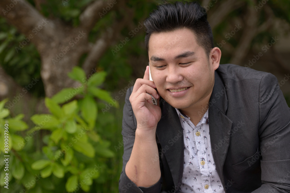 Happy young overweight Asian businessman talking on the phone at the park Happy young overweight Asian businessman talking on the phone at the park