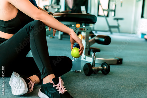 Woman exercise in gym fitness breaking and relax. hand holding apple fruit after training sport and dumbbell, water bottle on the ground.