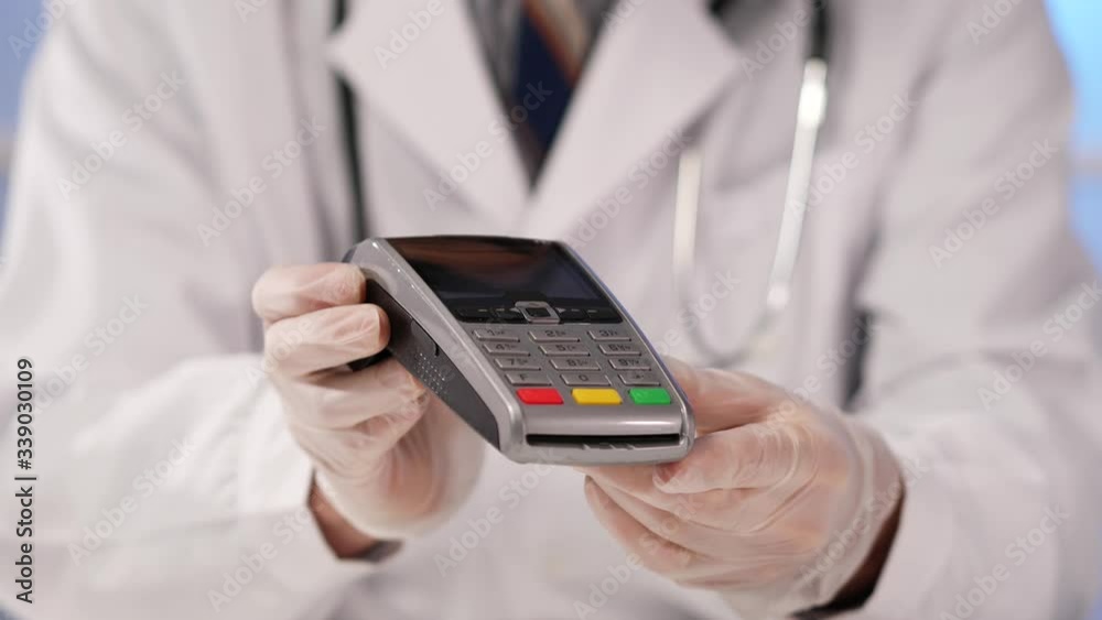 Closeup on a professional medical doctor, wearing a white lab coat and a stethoscope and protective gloves , holding a credit card terminal in his hands. Closeup on a professional medical doctor, wearing a white lab coat and a stethoscope and protective gloves , holding a credit card terminal in his hands.