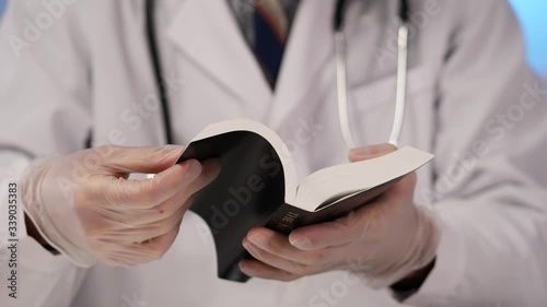 Medical professional doctor, wearing a white lab coat and a  stethoscope and protective gloves , holding and reading a Holy Bible closeup. photo