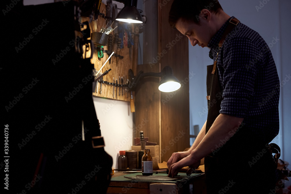 The leather master is working with natural leather at the desk. The room with wooden table and set of leather craft tools on it. Tools for craft production of leather goods on work desk. The leather master is working with natural leather at the desk. The room with wooden table and set of leather craft tools on it. Tools for craft production of leather goods on work desk.