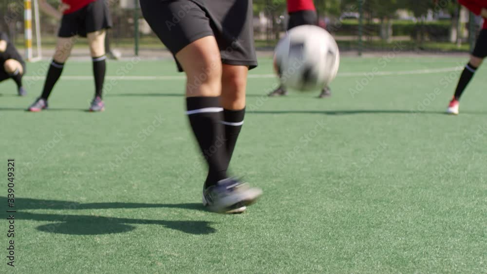 Low section shot of female soccer player juggling a ball on green field outdoors while teammates exercising in the background Low section shot of female soccer player juggling a ball on green field outdoors while teammates exercising in the background