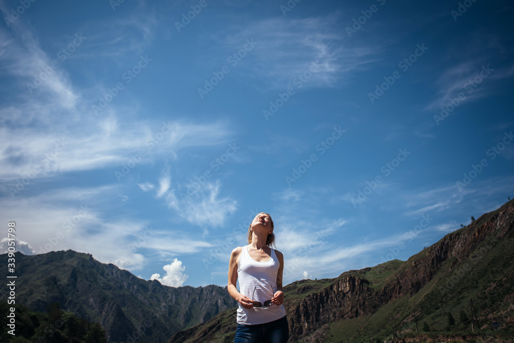 Naklejka premium Happy female traveller in a white t-shirt against the beautiful mountains and blue sky on sunny day. Cheerful young girl enjoying a trip in the Altai mountains.
