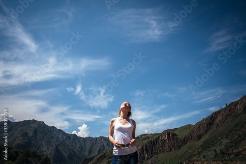 Happy female traveller in a white t-shirt against the beautiful mountains and blue sky on sunny day. Cheerful young girl enjoying a trip in the Altai mountains.