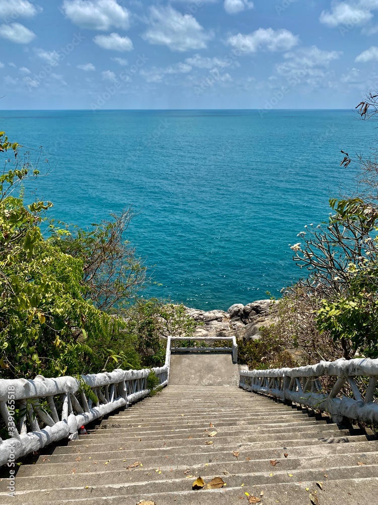 Fototapeta premium white staircase on the observation deck leading to the sea against a blue sky with white clouds and green trees under the sun in the tropics