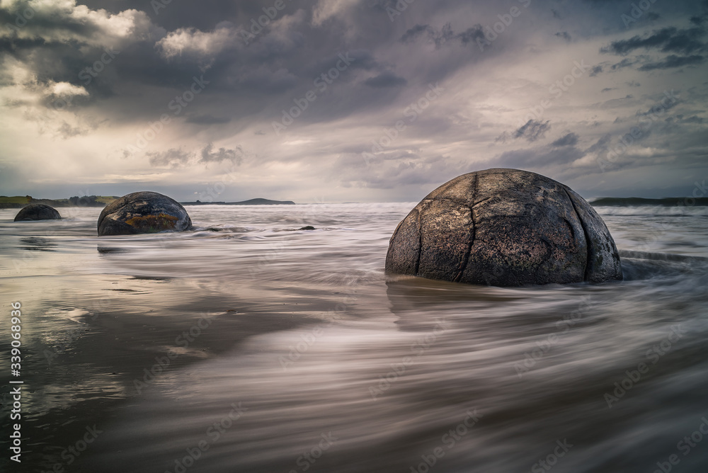 Obraz premium Waves washing against the mysterious Moeraki Boulders with dark storm clouds overhead at Koekohe beach on the Otago coast of New Zealand. These amazing round stones were formed millions of years ago
