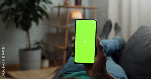 Young man lying on couch with modern smartphone in his hand while looking on mock up screen with trecking markers on it. Concept of green screen and chroma key. Cosy home background.