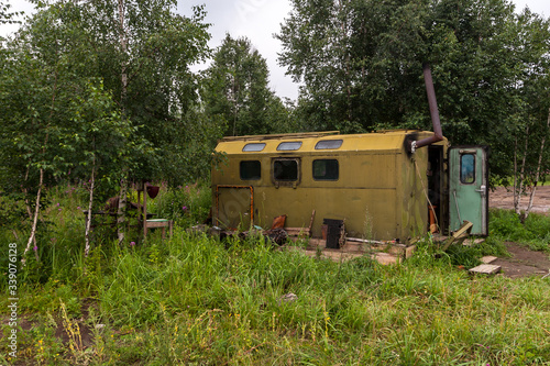 An old car booth converted into a house in the forest. Green iron shed with windows and chimney. The door is open. Around the green trees and grass. Horizontal.