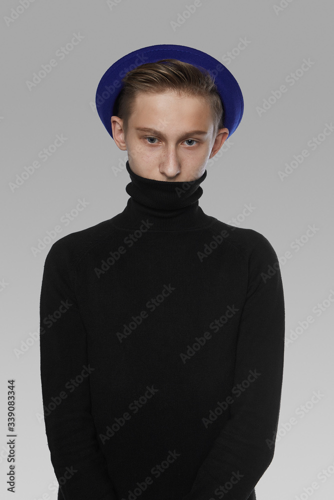 A young man with light brown hair is wearing a black turtleneck and a blue bowler hat. The boy's freckled face is half-hidden with the collar of his turtleneck. A young man with light brown hair is wearing a black turtleneck and a blue bowler hat. The boy's freckled face is half-hidden with the collar of his turtleneck.