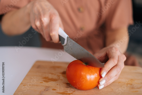 Close-up of hand of woman cutting fresh tomato using kitchen knife on wooden cutting board. Young woman cutting fresh organic tomato with a knife for vegetable salad.