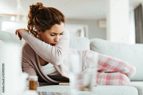 Sick woman sneezing into her elbow while lying down on sofa at home.