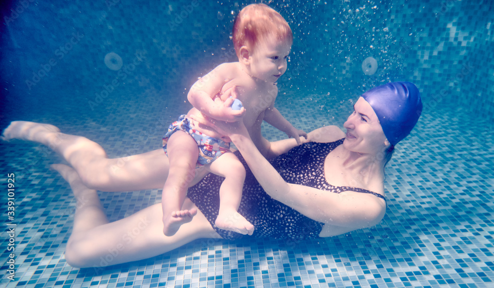 Smiling young woman in swimsuit holding adorable child in arms while lying on swimming pool floor underwater. Loving mother teaching son to swim underwater. Concept of family and swimming.