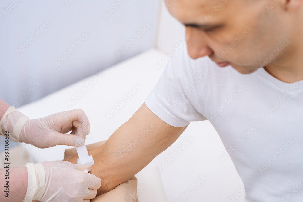 Fototapeta premium Nurse in rubber protective gloves putting an adhesive bandage on young man's arm vein after blood test or injection of vaccine, close-up. Concept of healthcare and medicine.