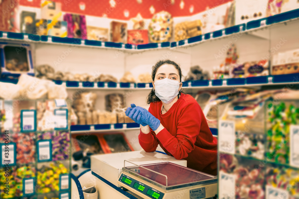 A bored young woman in gloves and a medical mask leaned against the counter,waiting for customers.In the background are shelves of the store.The concept of coronovirus and the crisis in business