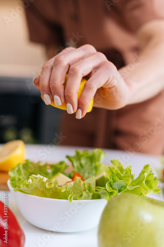 Close-up of women's hands squeezing juice from fresh yellow lemon into salad bowl with sliced vegetables. Red bell pepper, avocado, lemon on the table. Concept of healthy food lifestyle.