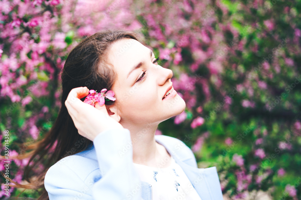 Portrait of young brunette girl with a flower behind her ear looks up at a blossoming pink apple tree and smiles. Close up. Portrait of young brunette girl with a flower behind her ear looks up at a blossoming pink apple tree and smiles. Close up.