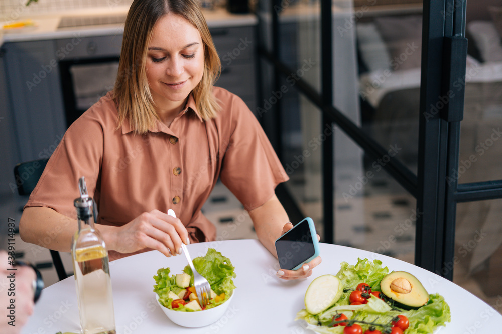 Young pretty woman mixing fresh vegetable salad while sitting at table in kitchen with modern interior, mobile phone on hand. Concept of healthy eating.