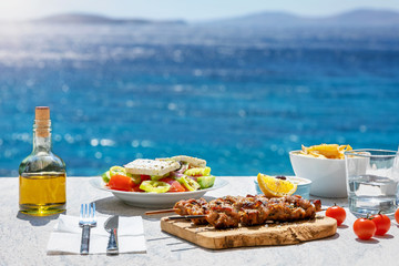 Greek food concept with farmers salad and souvlaki skewers in front of the sparkling, blue Aegean sea during summer time