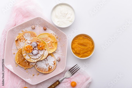 pancakes with raspberries and physalis, powdered sugar and maple syrup in pink plate with sour cream and mango jam on white background