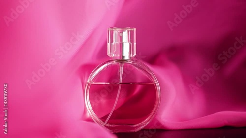 Oval surfaced bottle with pink perfumes or essential oils is on the table. Pink fabric flutters around and waves in the air and around the bottle. Concept of aroma and smell. Close up. Slow motion