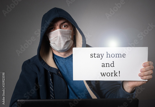 male hacker in a medical mask and a hood, works at a laptop and holds a tablet with the inscription - stay home and work photo