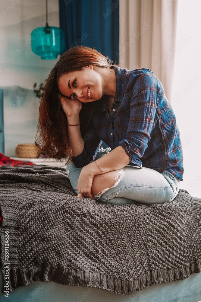 Fototapeta premium A brunette girl with long hair in a checked shirt is lying on the bed in a beautiful modern bright cozy interior