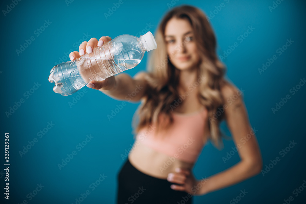 Attractive woman with blond hair in sport clothing standing in studio with bottle of water. Concept of refreshing and active lifestyle. Focus on bottle. Attractive woman with blond hair in sport clothing standing in studio with bottle of water. Concept of refreshing and active lifestyle. Focus on bottle.