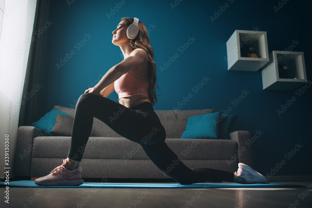 Fototapeta premium Young lady with muscular body in activewear exercising in headphones at home. Charming woman with blond hair stretching legs on yoga mat
