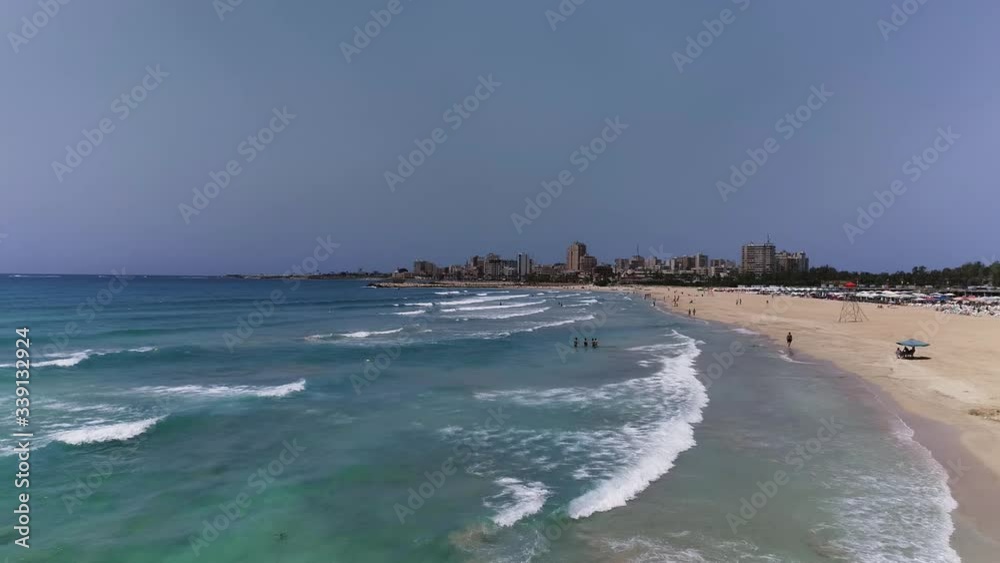 Aerial drone flying over crowded beach with ocean waves hitting the shore with city in the background Aerial drone flying over crowded beach with ocean waves hitting the shore with city in the background